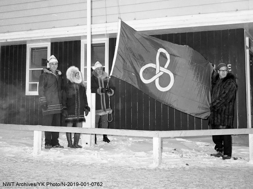 Quatre personnes en vêtements d’hiver déploient le drapeau des Jeux d’hiver de l’Arctique devant un bâtiment.