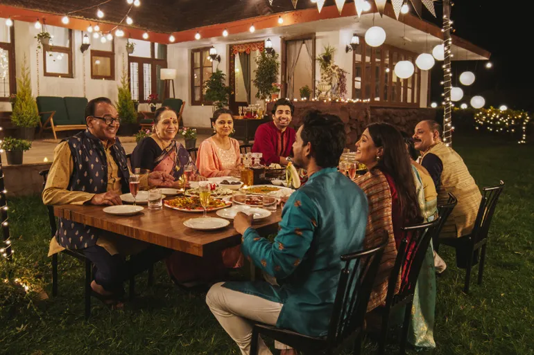 Une grande famille d’origine indienne partage un repas en soirée sur une terrasse décorée de guirlandes lumineuses.