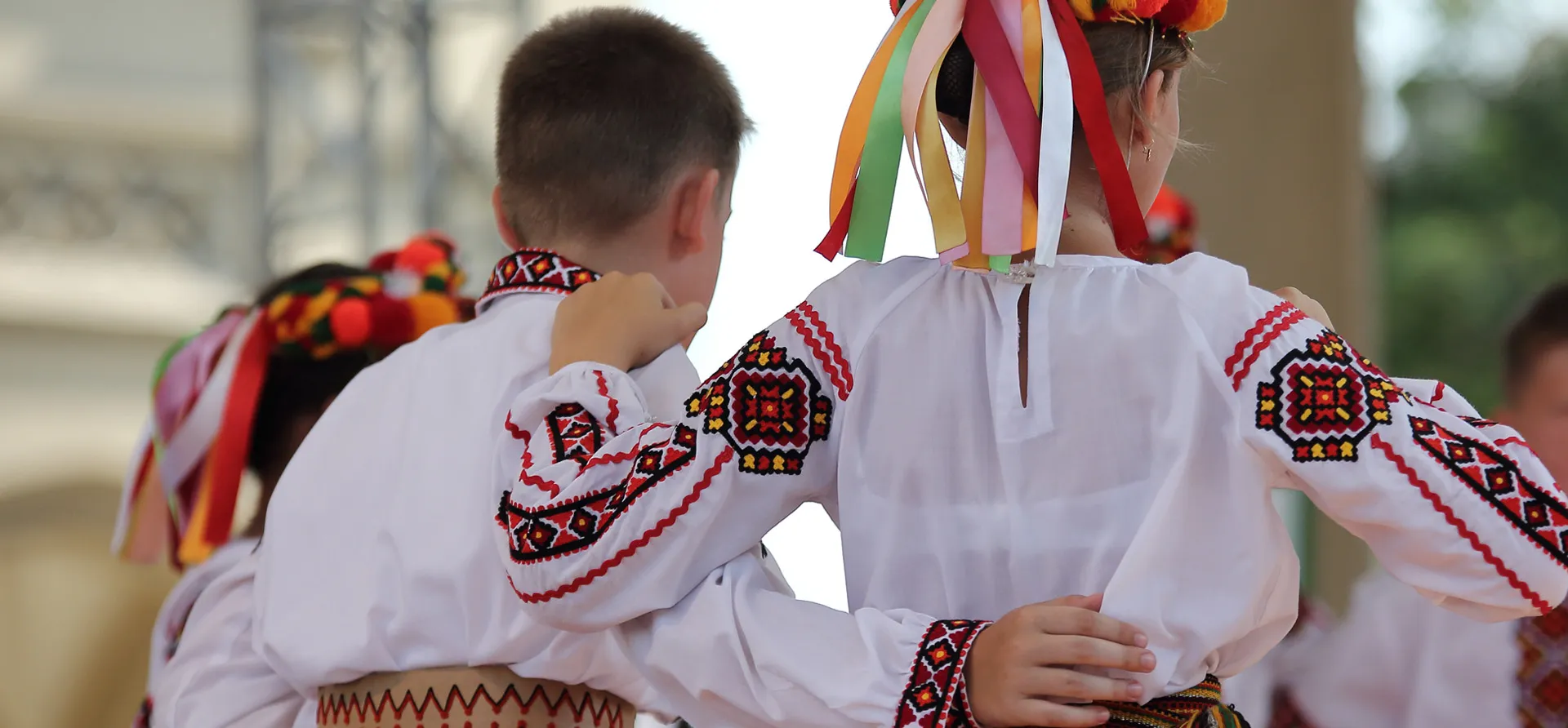 Un garçon et une fille vus de dos participent à une danse traditionnelle en chemises blanches brodées.
