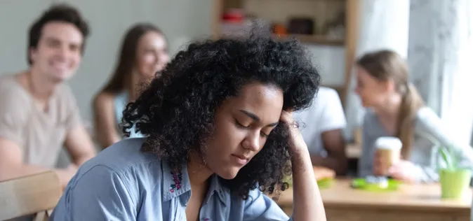 Une jeune fille triste a la tête baissée sur une table pendant que d’autres ados sourient derrière elle.