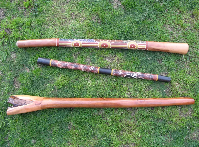 Trois didgeridoos en bois avec des motifs gravés sont posés par terre sur de l'herbe verte.